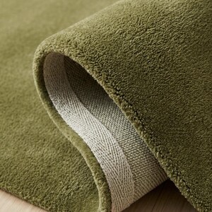 May include: A solid olive green area rug with a dense, short-pile texture. The rug is folded over to reveal a light beige woven backing along the edge, showcasing the thickness and construction of the textile material in a simple, clean view.