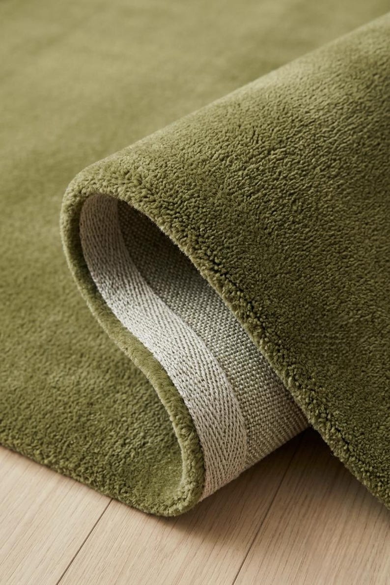 May include: A solid olive green area rug with a dense, short-pile texture. The rug is folded over to reveal a light beige woven backing along the edge, showcasing the thickness and construction of the textile material in a simple, clean view.