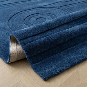 K&ouml;nnte beinhalten: Ein tiefblauer Teppich mit einer strukturierten Oberfl&auml;che, die geschnitzte konzentrische Kreise und parallele lineare Rillen aufweist. Der Teppich ist an einer Ecke umgeschlagen und zeigt ein hellbeiges Gewebe auf der R&uuml;ckseite.