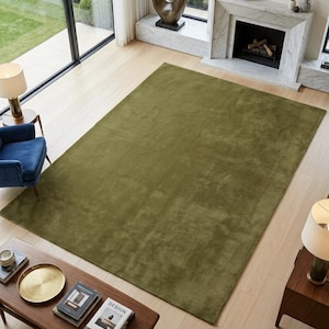 May include: A large, rectangular area rug in a solid olive green color with a smooth, low-pile texture. The rug is positioned centrally on a light-colored wooden floor, covering a significant portion of the floor space in the room.