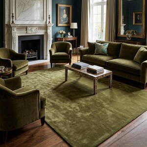 May include: A living room featuring a large olive green area rug, a matching velvet sofa, and three velvet armchairs. A rectangular coffee table with a patterned border sits in the center, while a marble fireplace and a gold-framed mirror are visible.