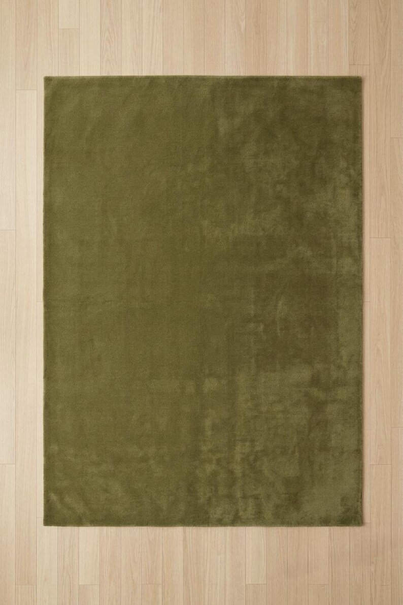May include: A rectangular area rug in a solid olive green color. The rug features a short, dense pile texture that creates subtle variations in light and shadow across its surface, providing a uniform appearance with clean, straight edges.