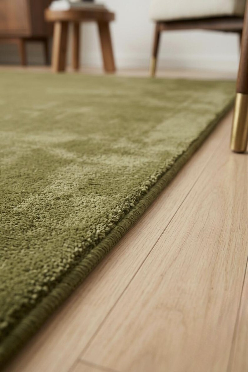 May include: A solid olive green area rug with a short, dense pile texture and a finished bound edge. The rug is rectangular in shape and features a consistent, uniform color throughout its surface, lying flat on a light-colored wooden floor.