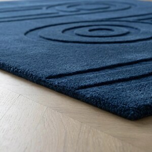 K&ouml;nnte beinhalten: Ein tiefblauer Teppich mit einer dicken, flauschigen Textur und einem geschnitzten, erhabenen Muster aus konzentrischen Kreisen und geraden parallelen Linien. Der Teppich weist eine einheitliche dunkelblaue Farbe auf seiner gesamten getufteten Oberfl&auml;che auf.