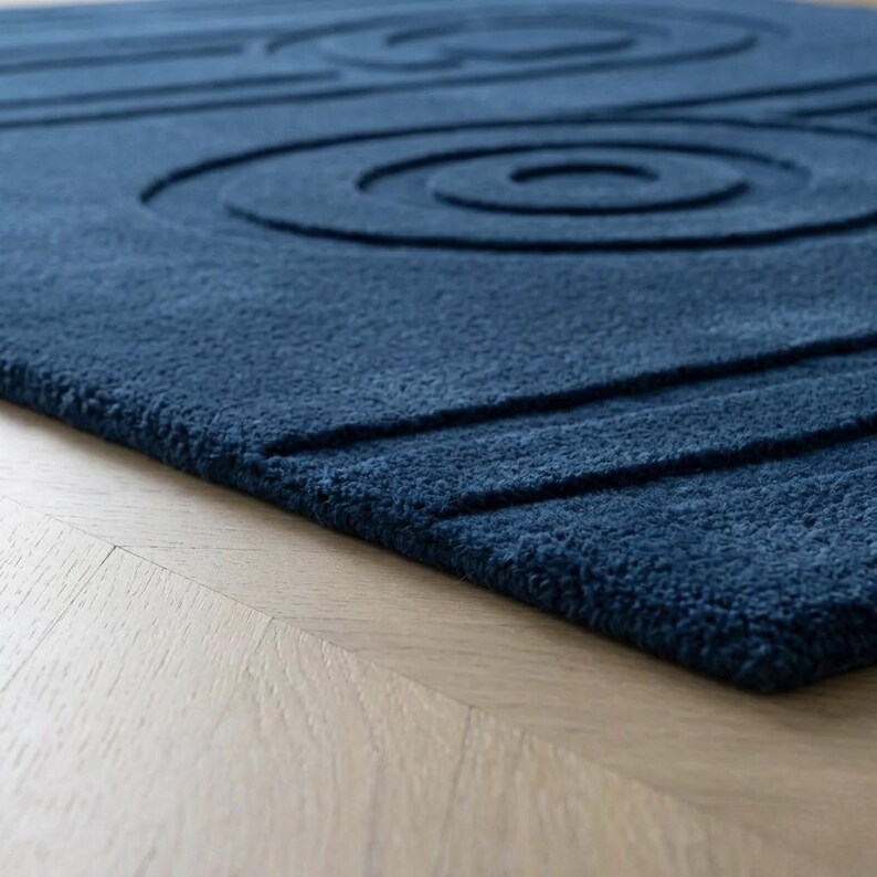 K&ouml;nnte beinhalten: Ein tiefblauer Teppich mit einer dicken, flauschigen Textur und einem geschnitzten, erhabenen Muster aus konzentrischen Kreisen und geraden parallelen Linien. Der Teppich weist eine einheitliche dunkelblaue Farbe auf seiner gesamten getufteten Oberfl&auml;che auf.