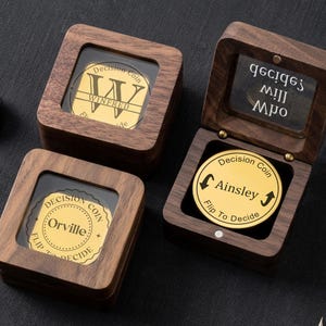 Peut inclure: Trois boîtes en bois contenant des pièces de décision. L'une est ouverte, révélant une pièce dorée avec le nom "Ainsley" et le texte "Flip To Decide". Les deux autres boîtes fermées affichent des pièces avec les noms "Winifred" et "Orville".