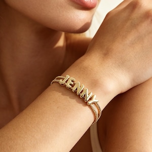 May include: A gold-toned bracelet featuring a double chain design and polished, rounded capital letters that spell out the name JENNY. The letters are centered on the wrist, creating a cohesive and metallic accessory with a smooth, reflective finish.