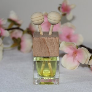 Può includere: Una bottiglia di vetro trasparente con un tappo di legno e due diffusori a forma di sfera di legno. La bottiglia contiene un liquido giallo. Fiori rosa e bianchi sullo sfondo.