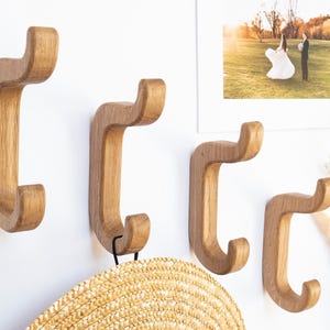 May include: Four wooden wall hooks with a curved design, mounted on a white wall. The hooks are a light brown colour and have a smooth finish. A straw hat hangs from one hook. A framed photo of a wedding is also visible.