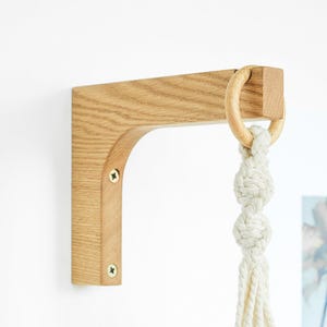 May include: A wooden plant hanger bracket with a natural finish. The bracket is mounted on a white wall and features a curved design. A wooden ring and knotted rope are attached for hanging a plant.