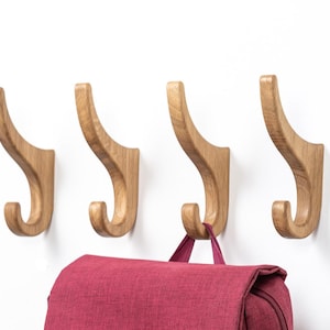 Op de afbeelding: Vier houten wandhaken met een gebogen ontwerp, gemonteerd op een witte muur. De haken zijn gemaakt van lichtgekleurd hout en hebben een gladde afwerking. Een rode rugzak hangt aan een van de haken.