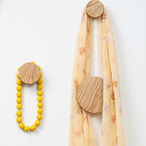 Op de afbeelding: Houten wandhaken met een cirkelvormig ontwerp. Eén haak houdt een gele kralenketting vast, terwijl een andere een lichtgele sjaal met een bloemenpatroon ondersteunt. De haken zijn op een witte muur gemonteerd en bieden een minimalistische esthetiek.