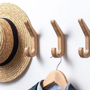 Peut inclure: Trois crochets muraux en bois de couleur naturelle sont fixés sur un mur blanc. Un chapeau de paille avec un ruban noir est suspendu à un crochet. Un cintre en bois avec une chemise est suspendu à un autre crochet. Les crochets ont un design simple et moderne.
