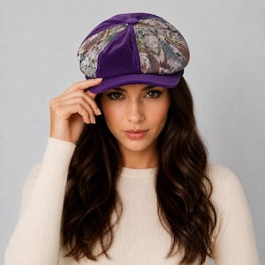 May include: A newsboy cap featuring a combination of solid purple corduroy panels and patterned fabric panels with a multi-colored abstract design. The hat has a rounded crown, a small button at the top center, and a short, stiff brim in purple.