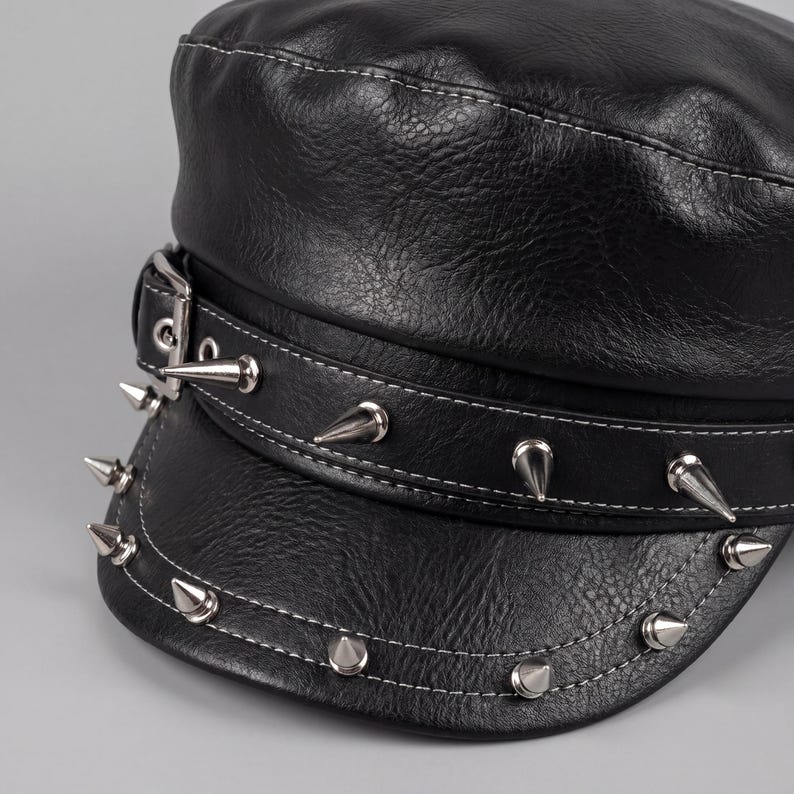 Puede incluir: Gorra de cuero negro con tachuelas c&oacute;nicas plateadas alrededor del borde y la banda. La gorra tiene un dise&ntilde;o estructurado con detalles de costura blanca. Una hebilla plateada asegura la banda.