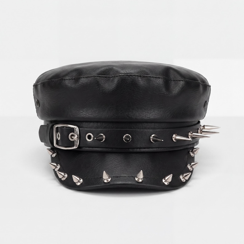 Puede incluir: Gorra negra de cuero sint&eacute;tico con la parte superior plana y una visera corta. El sombrero presenta una hebilla plateada y una banda adornada con p&uacute;as plateadas. P&uacute;as plateadas adicionales se colocan alrededor del borde.