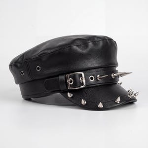 Puede incluir: Gorra de cuero negro con parte superior plana y visera corta. La gorra presenta un cintur&oacute;n con hebilla plateada y p&uacute;as plateadas alrededor de la visera y en el cintur&oacute;n. Estilo militar.