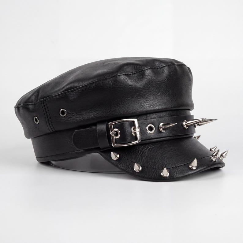 Puede incluir: Gorra de cuero negro con parte superior plana y visera corta. La gorra presenta un cintur&oacute;n con hebilla plateada y p&uacute;as plateadas alrededor de la visera y en el cintur&oacute;n. Estilo militar.