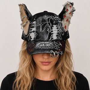 Puede incluir: Una gorra de béisbol de mezclilla negra con dos orejas grandes, deshilachadas y erguidas. La gorra está decorada con dibujos de líneas blancas, incluyendo un corazón y un ojo, patrones de punto de cruz rojo, anillos de metal plateado y parches.