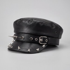 Puede incluir: Gorra negra de cuero sint&eacute;tico con p&uacute;as de metal plateado y cierre de hebilla. El sombrero tiene una parte superior plana y una visera corta, con una correa decorativa. Un accesorio de moda.