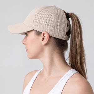 May include: A beige baseball cap with a curved peak and a perforated crown. The cap has a ponytail opening at the back. The cap is worn by a person wearing a white vest top.
