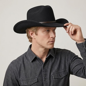 May include: A black felt cowboy hat with a wide, curved brim and a thin, dark brown leather band around the crown. The hat features a structured, pinched crown design and is worn with a dark grey, long-sleeved button-down shirt with two pockets.