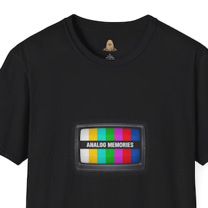 May include: Black t-shirt with a graphic of a retro television screen. The screen shows a colourful test pattern with the text "ANALOG MEMORIES". The t-shirt has a crew neck and short sleeves, a classic design.
