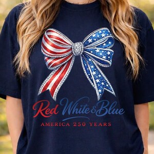 May include: Dark blue t-shirt with a bow design in red, white, and blue, with a star pattern. The text reads "Red White & Blue AMERICA 250 YEARS". A patriotic themed top.
