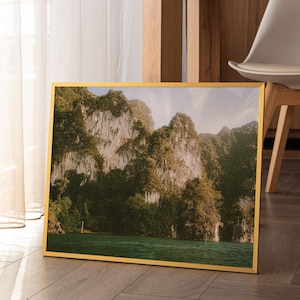 May include: A framed landscape photograph depicting a mountain range with green vegetation and a body of water. The image is set in a gold frame and leans against a wall. The scene is illuminated by natural light.