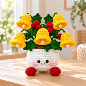 May include: A crocheted white pot with a smiling face, pink cheeks, and two red feet, containing a bouquet of green holly leaves, red berries, and five bright yellow crocheted bells, each featuring a small white pearl bead at the center.