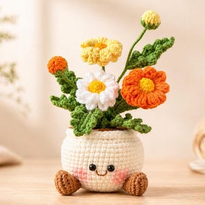 May include: A crocheted flower arrangement in a white pot with a smiling face and small brown feet. The arrangement includes orange, yellow, and white flowers with green leaves. The pot is sitting on a wooden surface.