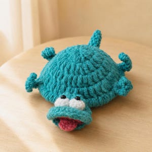 May include: A teal crocheted turtle-like creature with a rounded shell, four small legs, and a tail. The head features two white circular eyes with black pupils, a wide mouth opening, and a small pink tongue protruding from the center.