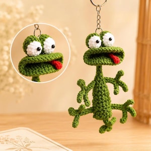 May include: A green crocheted frog keychain with white eyes and a red tongue. The frog has a round head, a long neck, and four limbs. The keychain is attached to a silver metal ring.