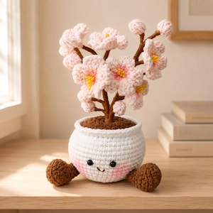 May include: A crocheted cherry blossom tree in a white pot with a smiling face, pink cheeks, and two brown feet. The tree has brown branches and light pink flowers with yellow centers. The pot is textured with white yarn and has a brown soil top.