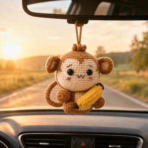 May include: A crocheted monkey ornament hanging from a rearview mirror. The monkey is light brown and cream with large black eyes, a small stitched mouth, and pink blush marks. It holds a bright yellow banana in its arms and has a curled tail.