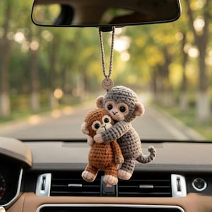 May include: A crochet car hanging ornament featuring two small monkeys hugging. One monkey is light brown with a cream face and belly, while the larger monkey is grey with a cream face. They are suspended by a beige cord with a small smiley face charm.
