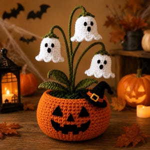 May include: A crocheted orange jack-o'-lantern pot containing three white ghost-shaped flowers with green stems and leaves. A small black witch hat with an orange band sits on the rim of the pumpkin pot, which features a black stitched face.