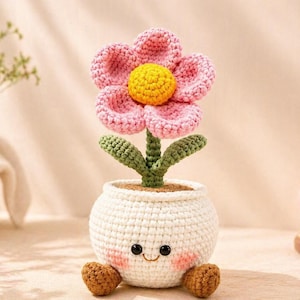 May include: A crocheted flower in a white pot with a smiling face. The flower has pink petals and a yellow center. The pot has brown feet and pink cheeks. The flower is a handmade item.