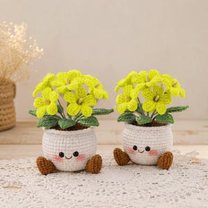 Puede incluir: Dos macetas de flores de ganchillo con caras sonrientes, pies marrones y flores amarillas con hojas verdes. Las macetas blancas están sobre un mantel individual de encaje blanco, creando una exhibición encantadora.