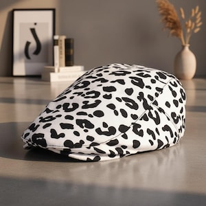 May include: A white and black patterned newsboy cap. The cap features a repeating pattern of black spots on a white background. The cap is made of a soft material and has a curved brim.
