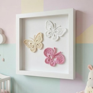 May include: A white framed wall art piece featuring three crocheted butterflies in cream, white, and pink. The butterflies are arranged on a white background, creating a delicate and whimsical aesthetic. The frame is square and painted white.