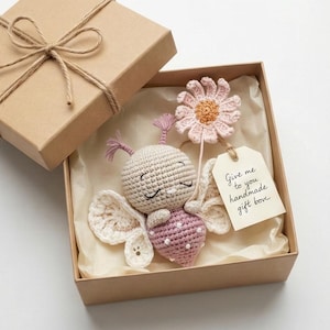 Puede incluir: Un conjunto de mariposa y flor de ganchillo hechos a mano en una caja de regalo. La mariposa es beige y rosa con lunares blancos. La flor es rosa y naranja. La etiqueta dice "Give me to you handmade gift box".