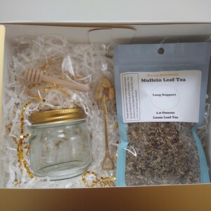 May include: A gift box containing a clear glass jar with a gold lid, a wooden honey dipper, a gold-toned shovel-shaped spoon, and a 1.0 ounce bag of Mullein Leaf Tea labeled Lung Support, all nestled in white and gold shredded paper filler.