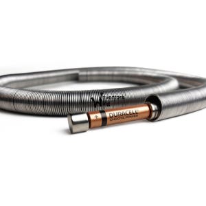 Könnte beinhalten: Ein silberfarbenes, flexibles Metallrohr mit einer Duracell-Batterie und einer silbernen Kappe. Die Batterie ist kupferfarben mit schwarzem Text "DURACELL CHHOTA POWER". Das Rohr ist auf weißem Hintergrund gewickelt.