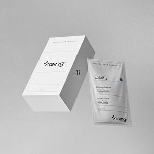 Op de afbeelding: Een witte doos en een zakje met het woord "rising" in een gestileerd lettertype. Op de doos staat de tekst "ENJOY YOUR POTENTIAL" en op het zakje staat "Clarity" met een blauwe stip. Het zakje vermeldt "Concentratie, Productiviteit" en andere details.