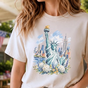 Puede incluir: Camiseta beige con una ilustraci&oacute;n en acuarela de la Estatua de la Libertad y el horizonte de la ciudad de Nueva York. El dise&ntilde;o incluye detalles florales azules, blancos y amarillos. La camiseta tiene mangas cortas.