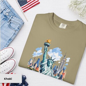 Puede incluir: Camiseta color caqui con una ilustraci&oacute;n en acuarela de la Estatua de la Libertad y el horizonte de la ciudad de Nueva York. La camiseta se muestra con vaqueros azules, zapatillas blancas y una peque&ntilde;a bandera americana.