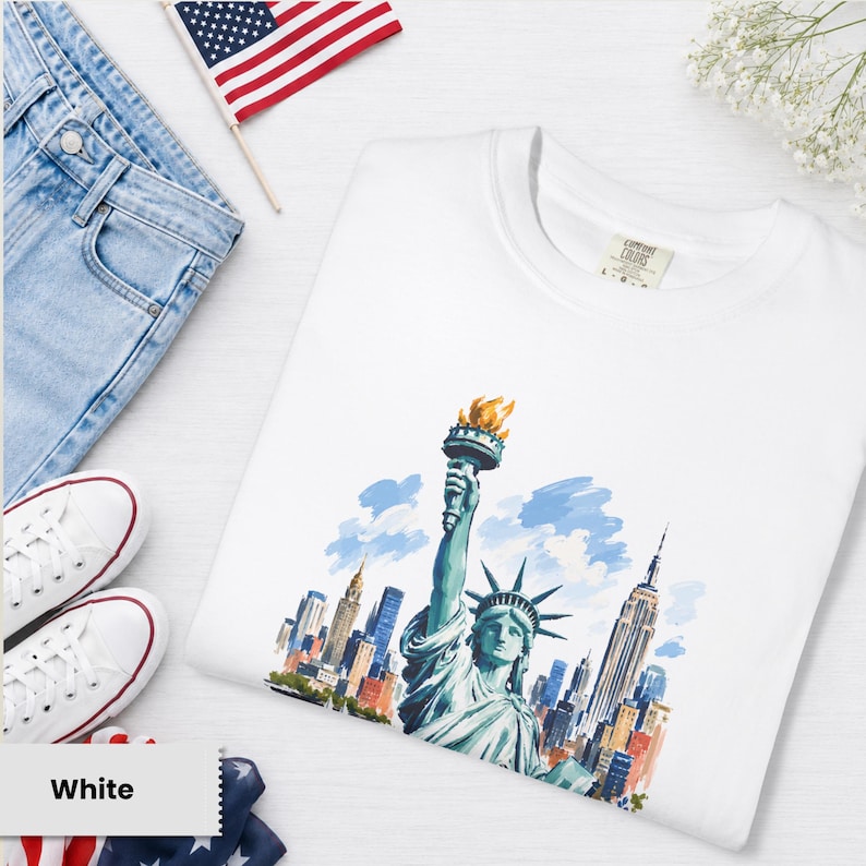 Puede incluir: Camiseta blanca con una ilustraci&oacute;n en acuarela de la Estatua de la Libertad y el horizonte de la ciudad de Nueva York. La camiseta se muestra con vaqueros azules, zapatillas blancas y una peque&ntilde;a bandera americana.