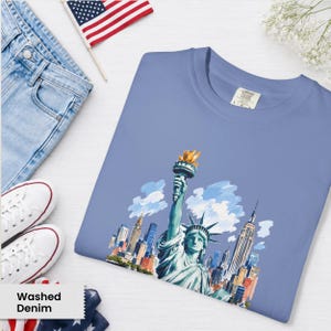 Puede incluir: Una camiseta azul claro con una ilustraci&oacute;n en acuarela de la Estatua de la Libertad y el horizonte de la ciudad de Nueva York. La camiseta se muestra con vaqueros de lavado claro, zapatillas blancas y una peque&ntilde;a bandera americana.