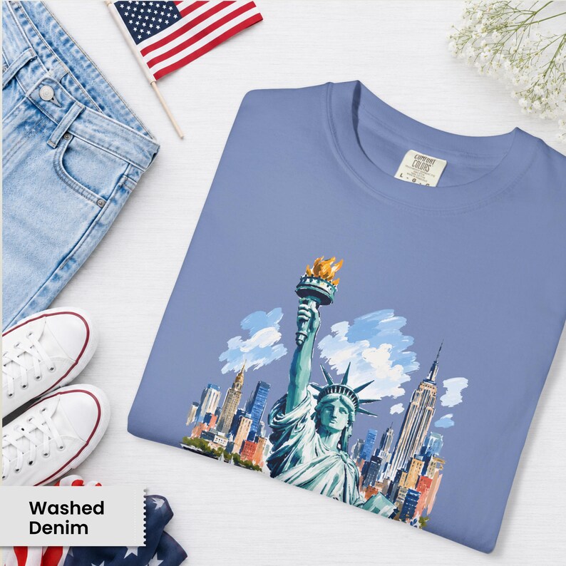 Puede incluir: Una camiseta azul claro con una ilustraci&oacute;n en acuarela de la Estatua de la Libertad y el horizonte de la ciudad de Nueva York. La camiseta se muestra con vaqueros de lavado claro, zapatillas blancas y una peque&ntilde;a bandera americana.
